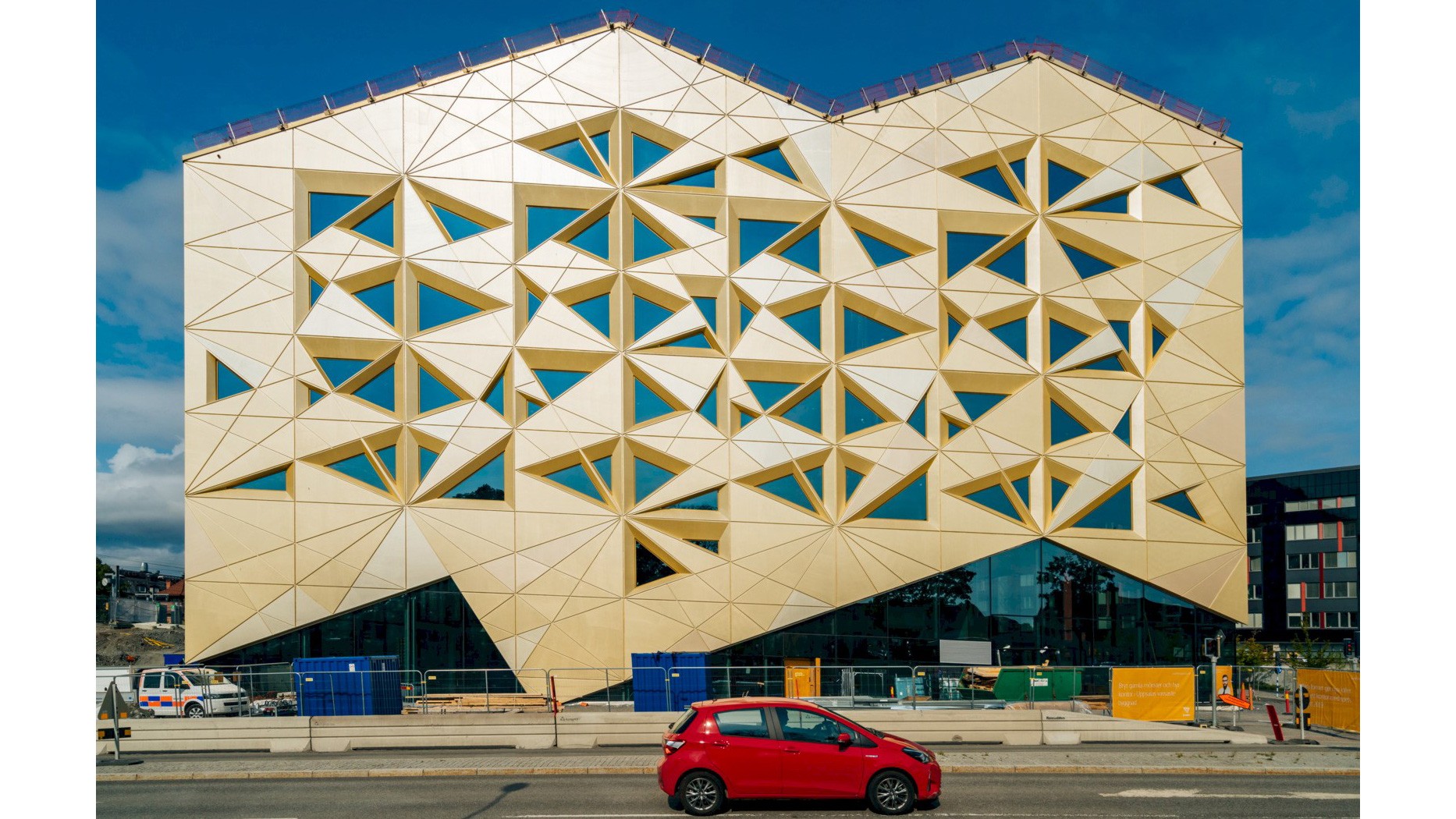 Modern byggnad med geometrisk fasad i livlig guld, med triangulära fönster; röd bil passerar.