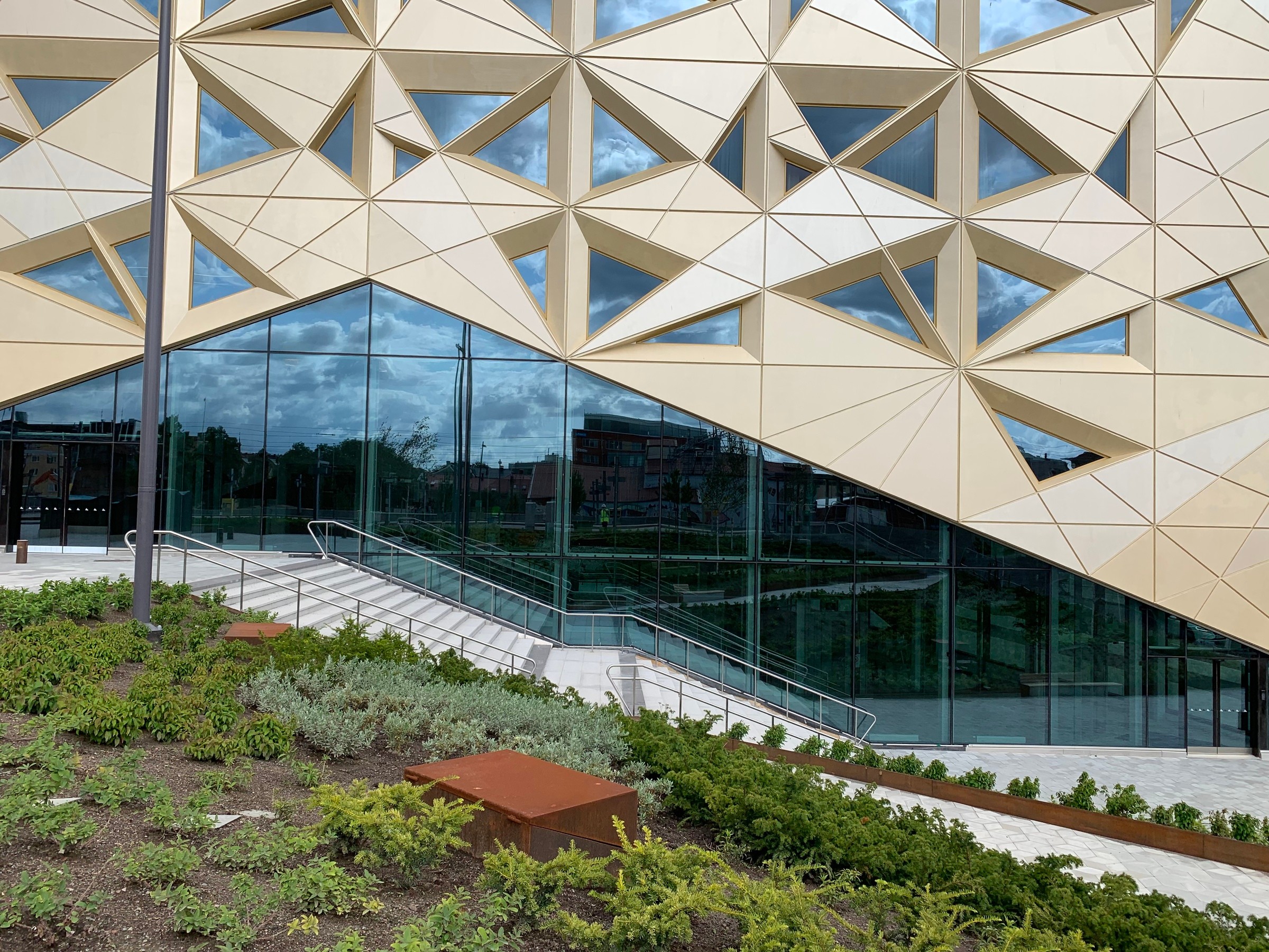 Modern byggnad med geometrisk fasad, inglasad entré och anlagd grönska i förgrunden.