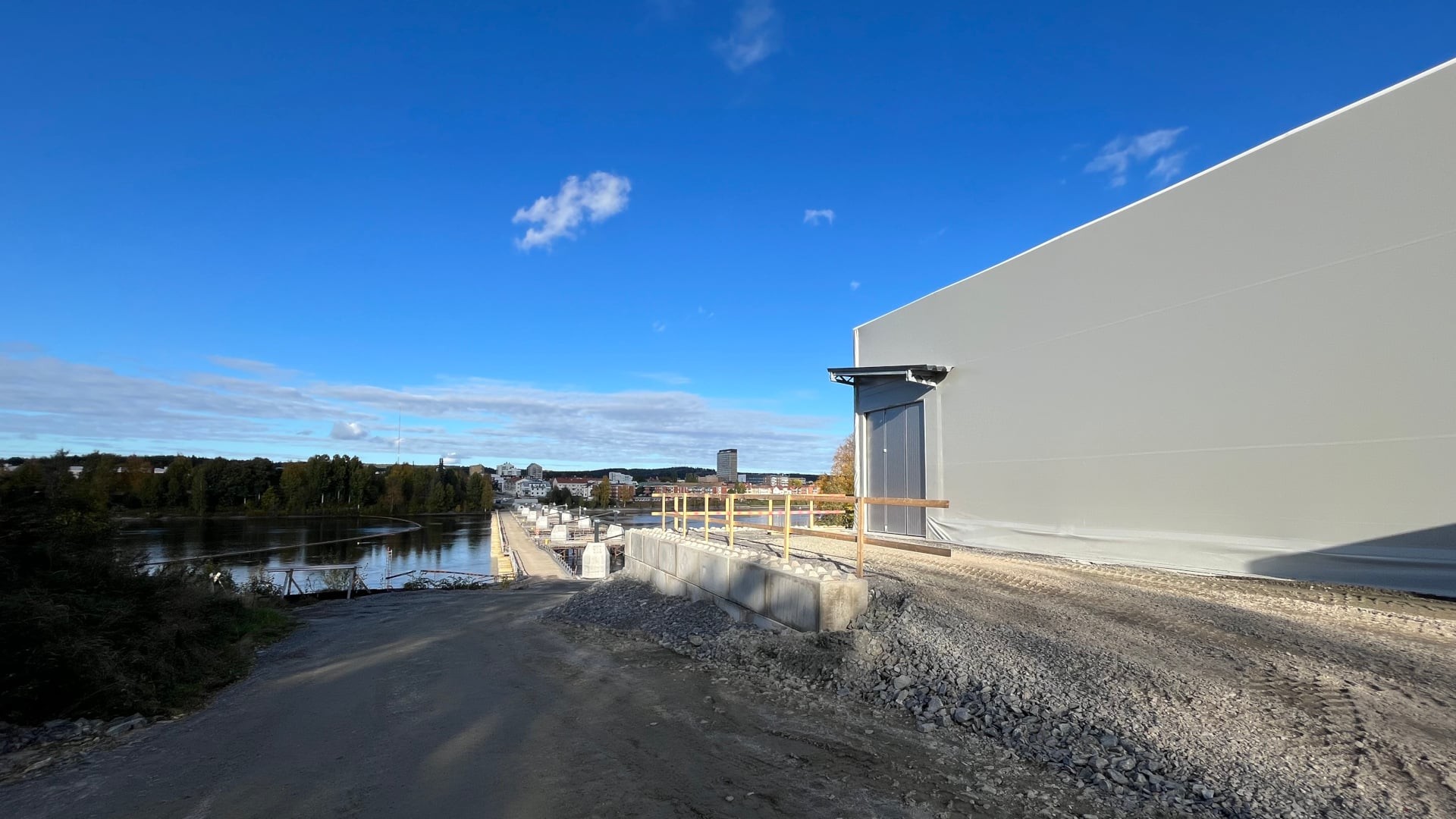 Stort väderskydd vid Skellefteälvens strand där Karlgårdsbron monteras, med stadssilhuetten i bakgrunden under klarblå himmel.