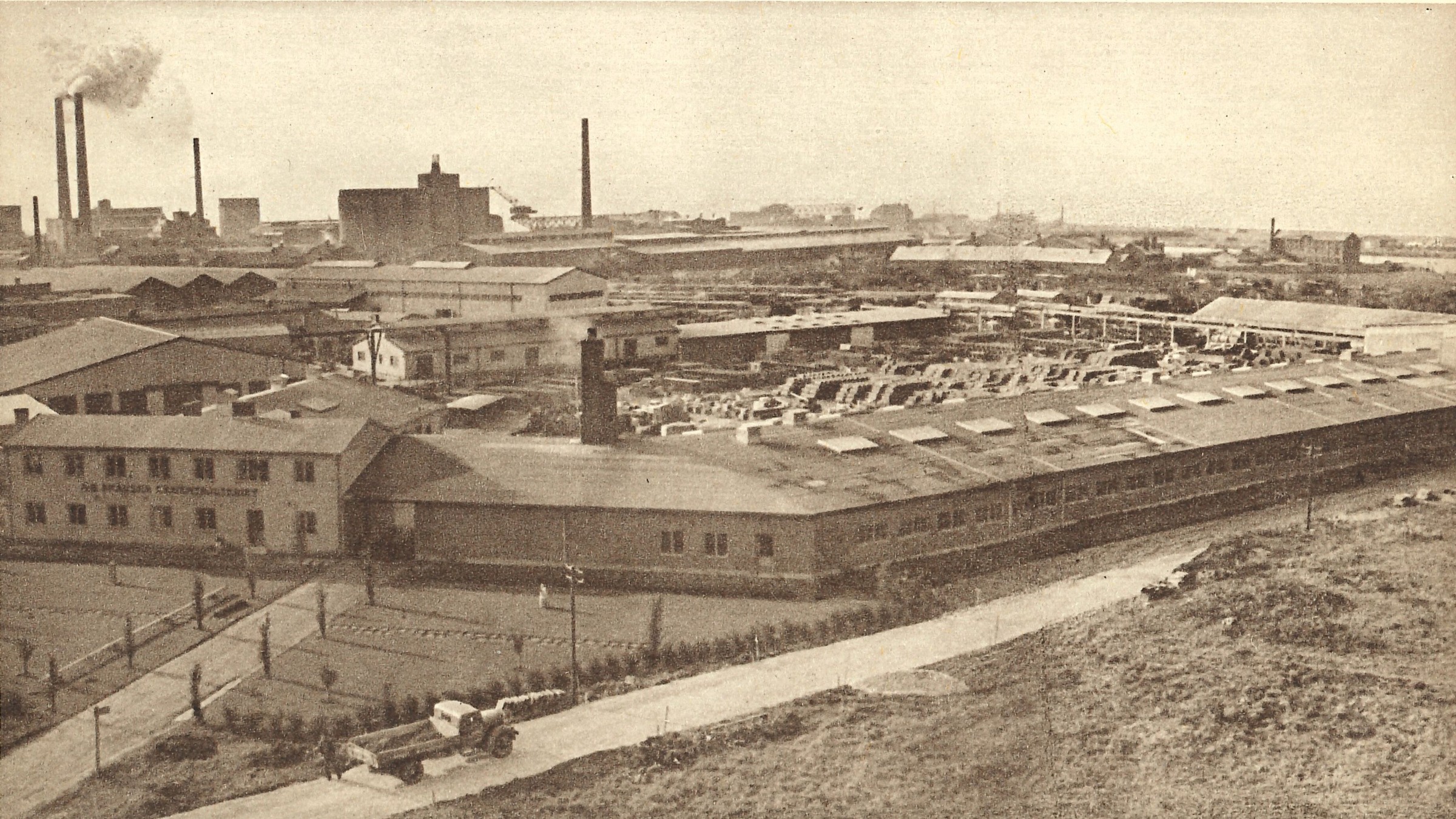 En flygvy av ett industrikomplex från mitten av 1900-talet med fabriker och skorstenar i bakgrunden.