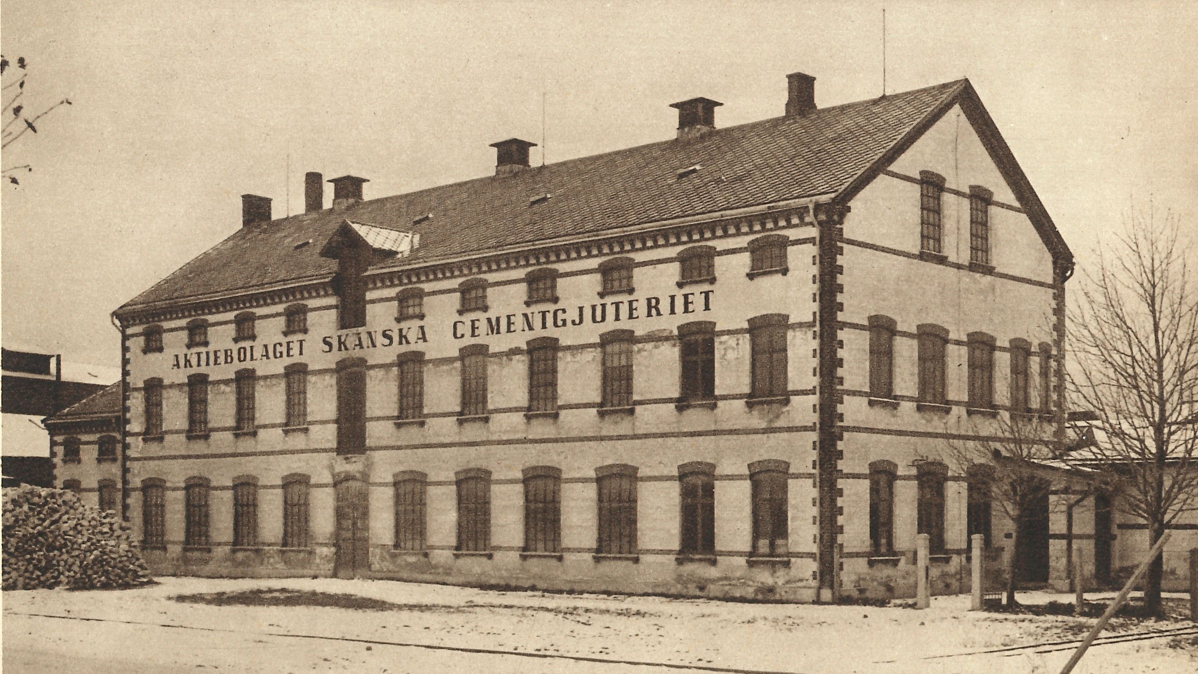 Historisk byggnad med skylten "Aktiebolaget Skånska Cementgjuteriet", snötäckt mark, gammalt foto.