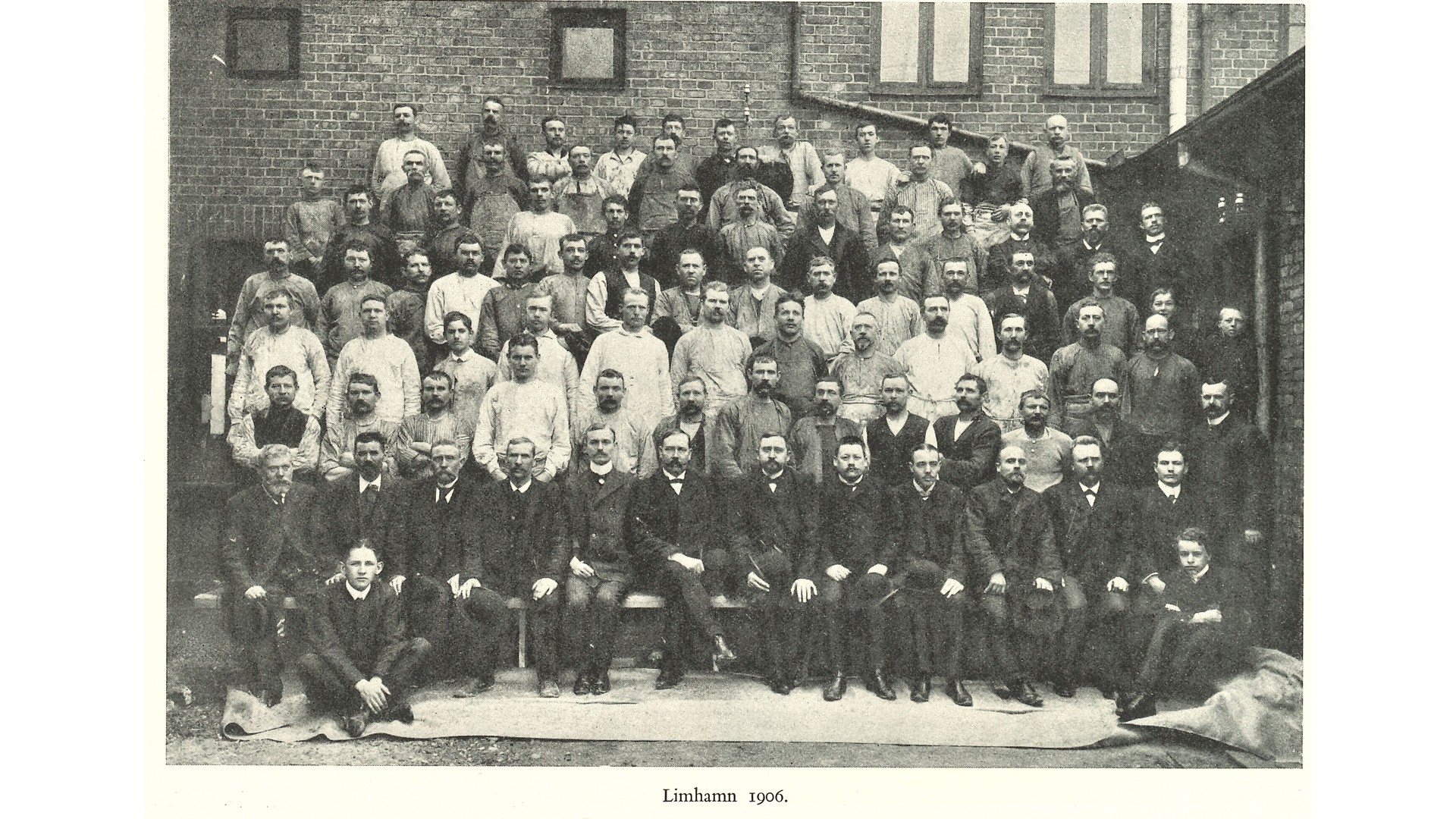 Gruppfoto av män i formell och arbetsklädsel i Limhamn, 1906, med en tegelbyggnad som bakgrund.