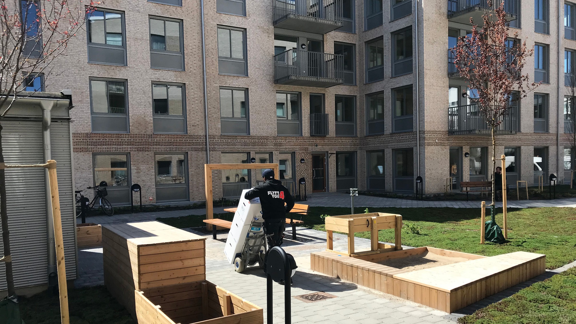 Modern innergård i lägenhetskomplex med trälådor för plantering, bänkar och en person som arbetar.