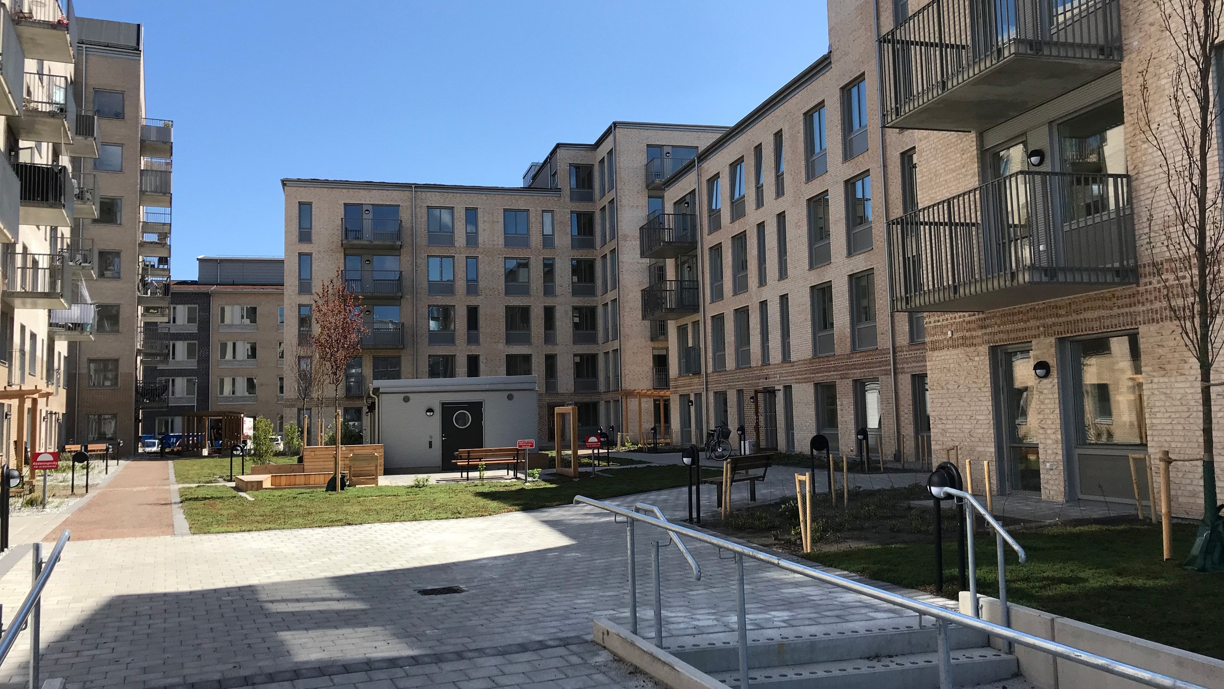 Modernt lägenhetskomplex med flera balkonger, anlagd innergård och klarblå himmel.