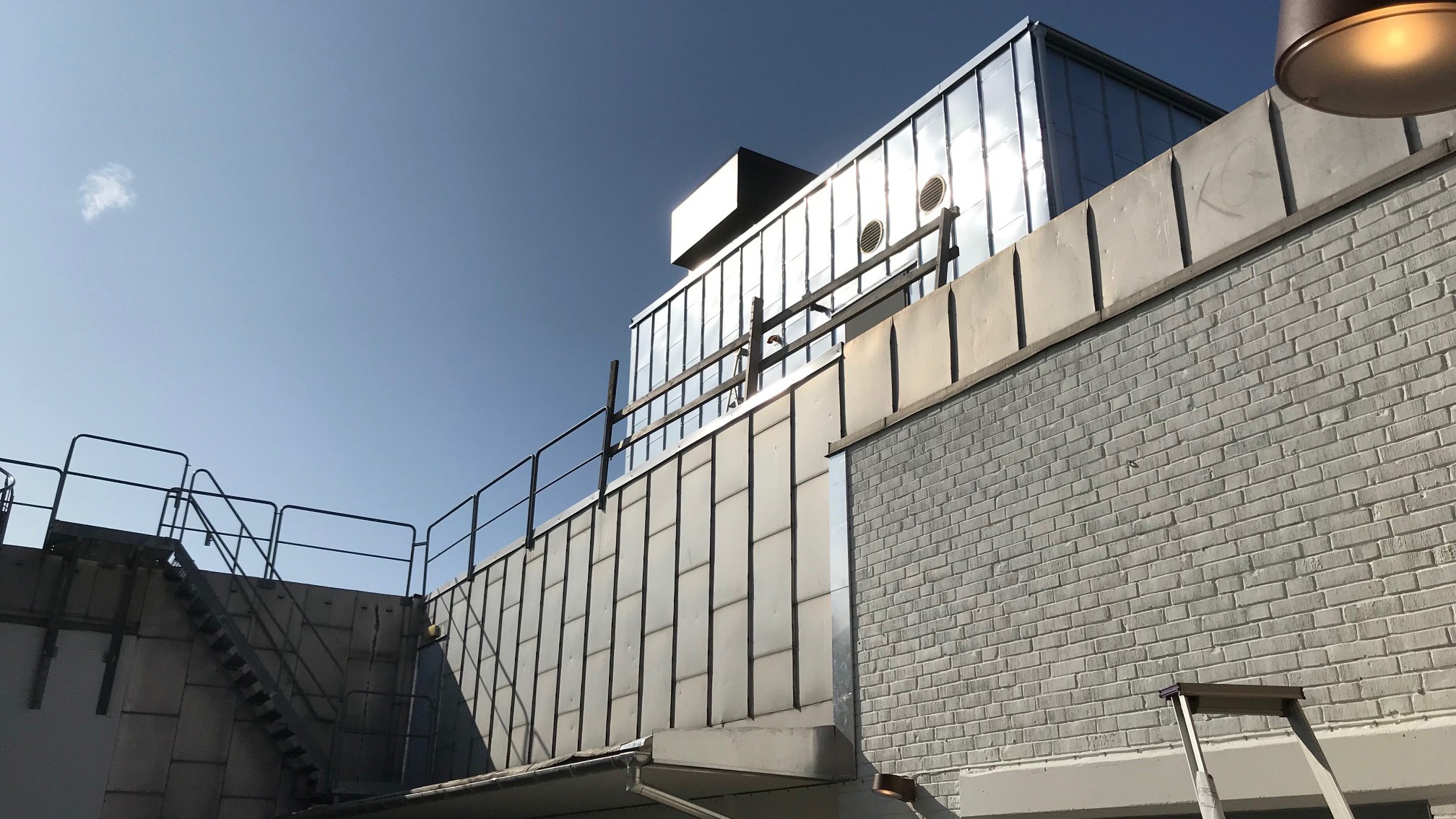 Industriellt tak med metallbeklädnad, räcken och en trappa under en klarblå himmel.