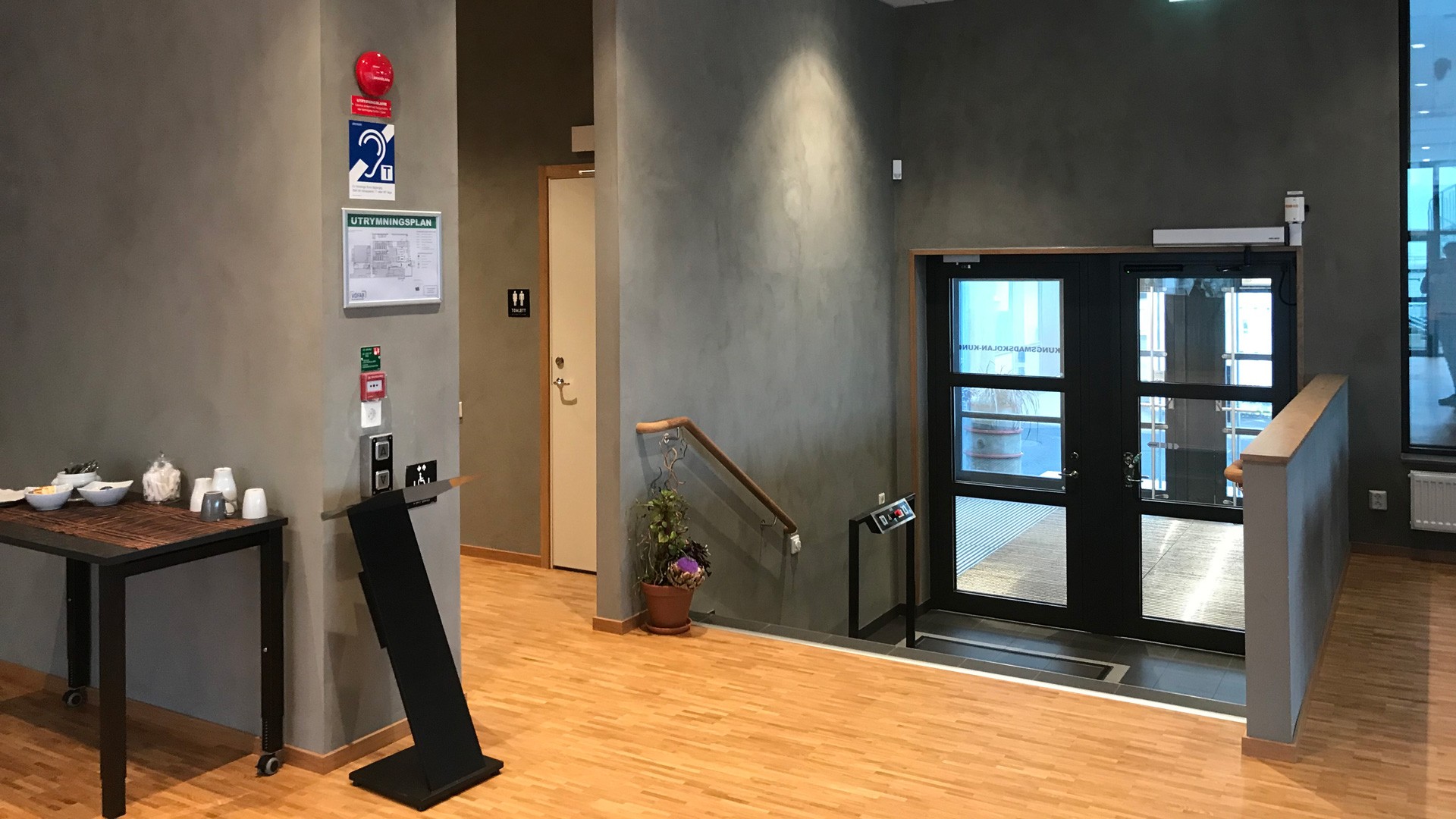 Modern lobby med trägolv, informationsskyltar och en trappa som leder till glasdörrar.