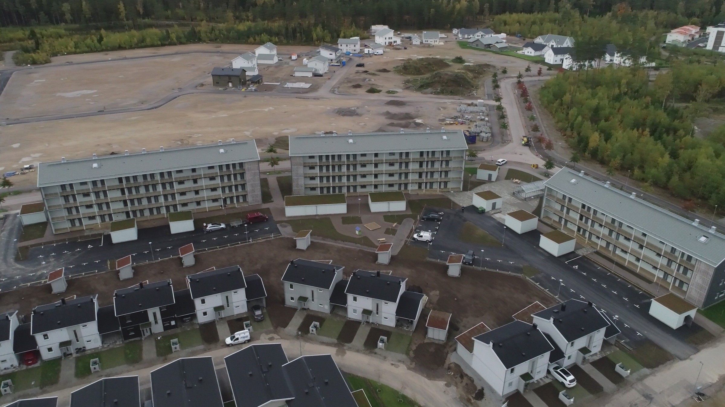 Flygvy över ett bostadsområde med moderna lägenhetsbyggnader och hus nära en skog.