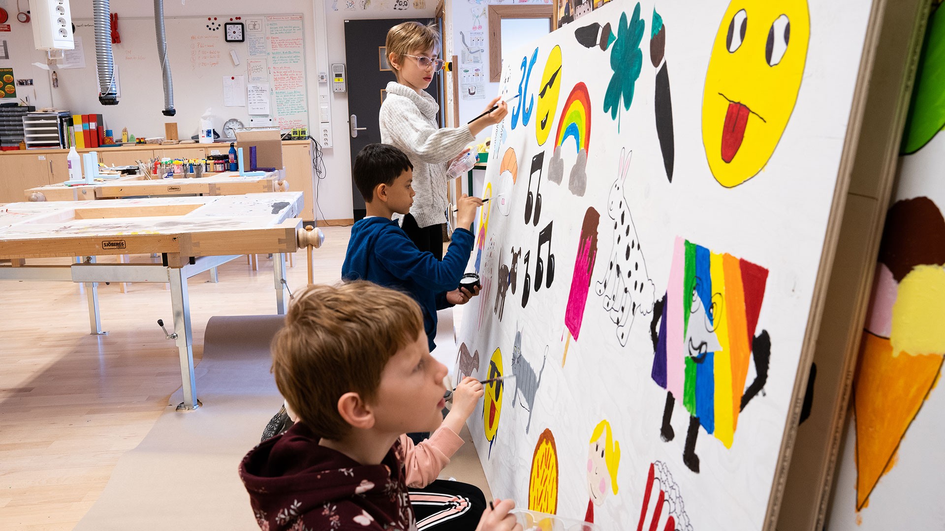 Barnen har inte målat i så stort format tidigare och fick börja med att rita upp sina figurer i liten skala på papper och sedan förstora dem på plywoodskivorna. Barn målar en färgglad väggmålning i ett klassrum, med emojis och regnbågsdesign.