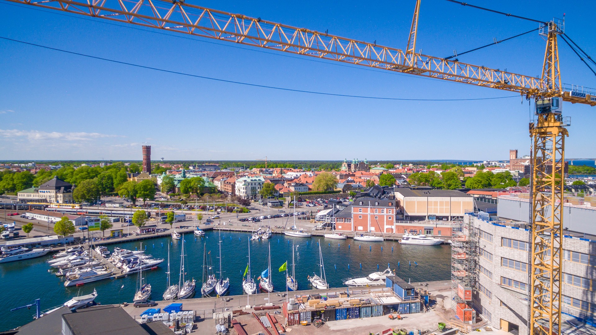 Flygvy över en livlig hamn med båtar förtöjda, omgiven av byggnader och en byggkran.