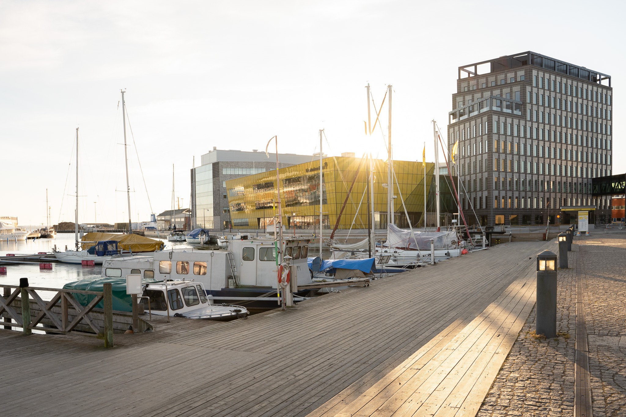 Modern hamnmiljö med förtöjda båtar, träbrygga och solbelysta samtida byggnader.