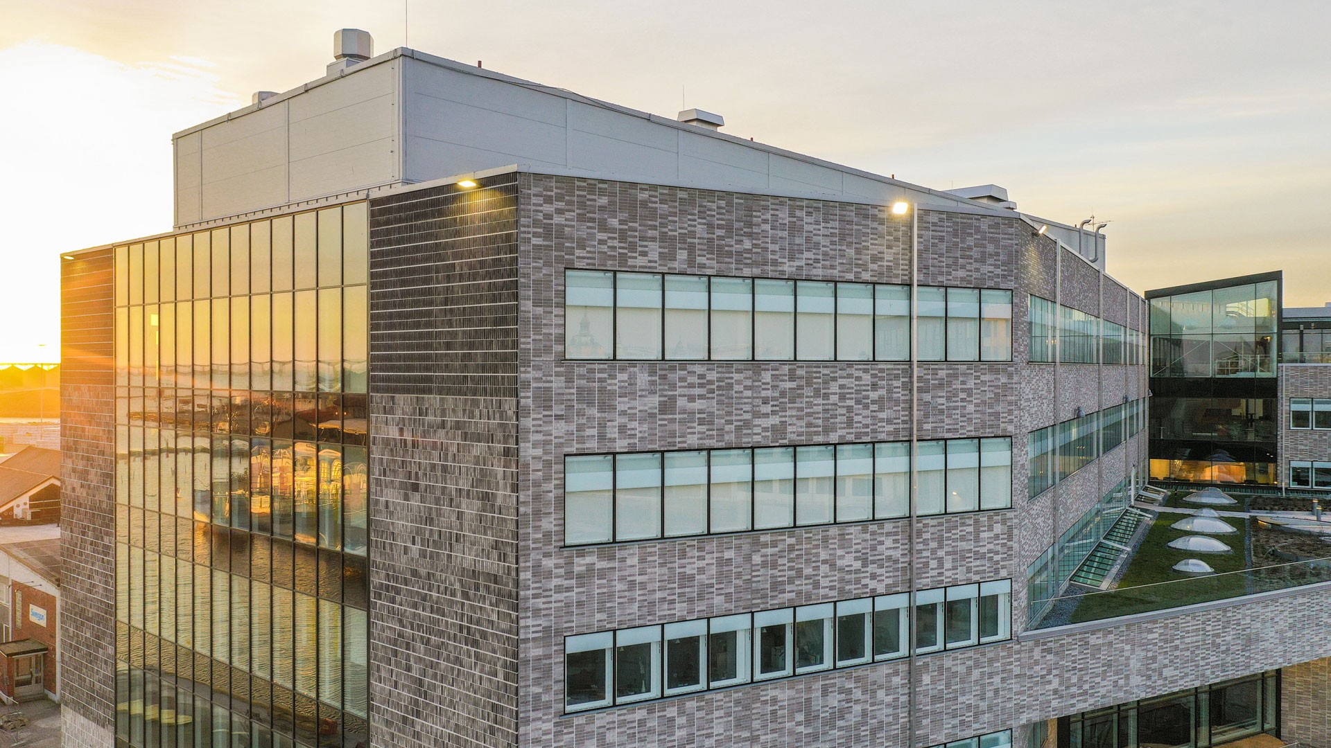 Modern tegelkontorsbyggnad med stora fönster som reflekterar solnedgången, med en takterrass.