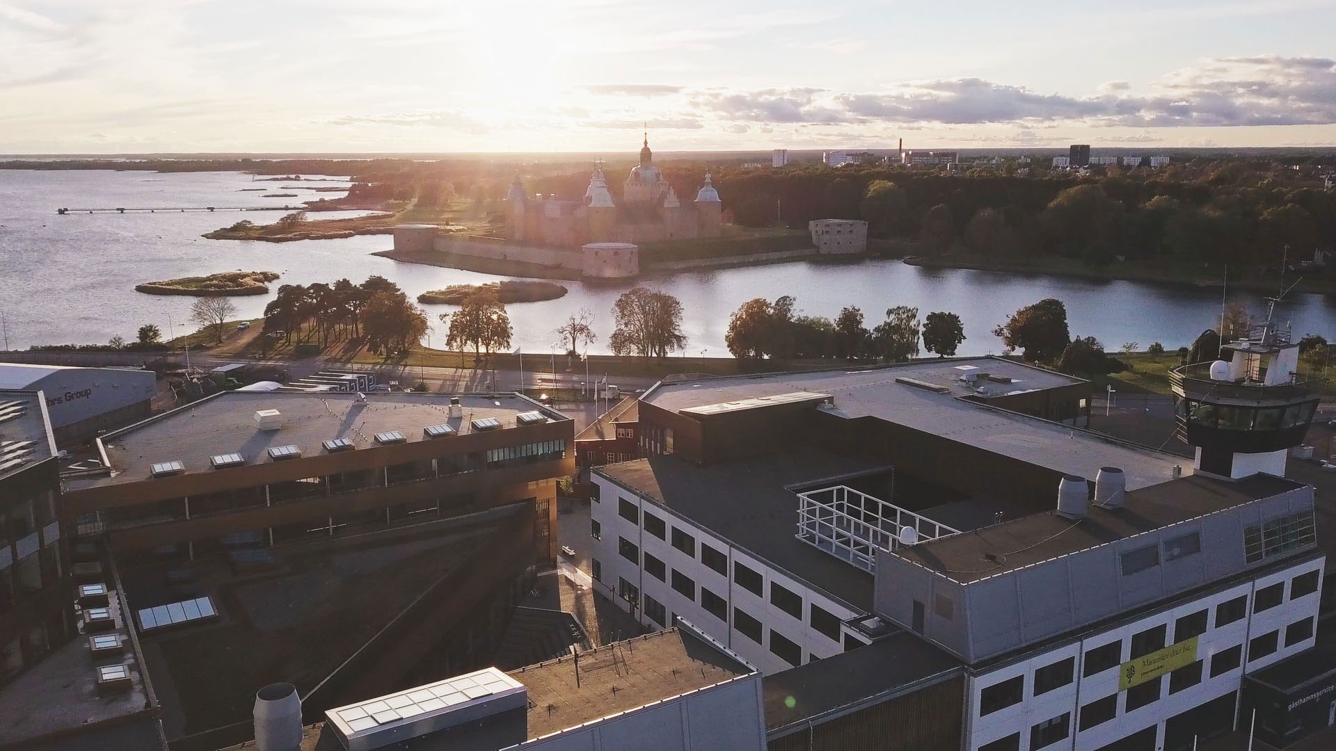 Flygvy över ett naturskönt slott vid en sjö vid solnedgången, omgiven av moderna byggnader och frodiga träd.