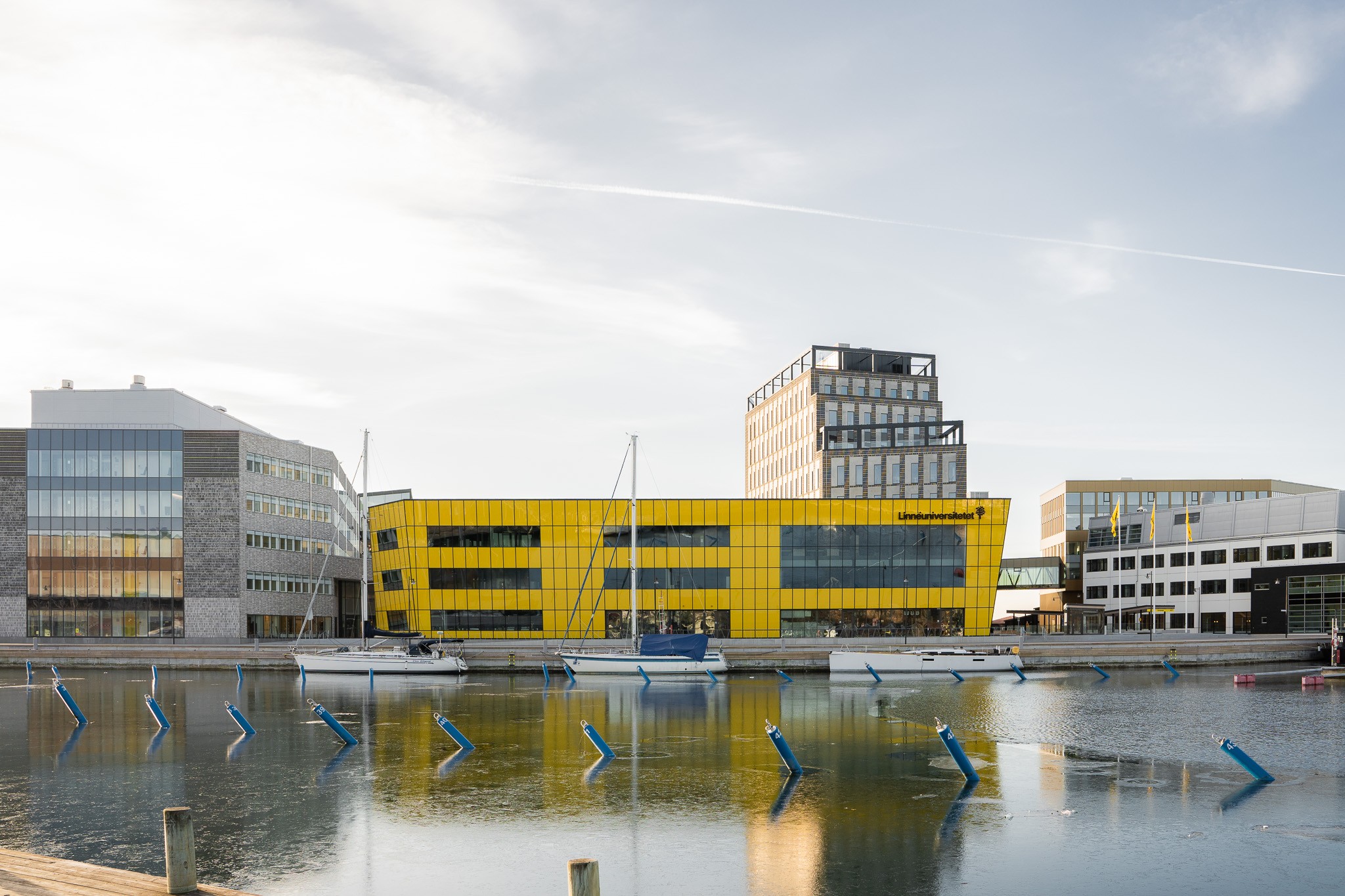 Skanska fick i uppdrag att hyra ut, bygga nytt och bygga om vid Linnéuniversitet i Kalmar. Moderna byggnader vid vattnet med en framträdande gul struktur som speglas i lugnt vatten.