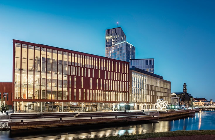 Malmö Live Modern arkitektonisk byggnad med glasfasad som vetter mot en vattenväg i skymningen i en stadsomgivning.