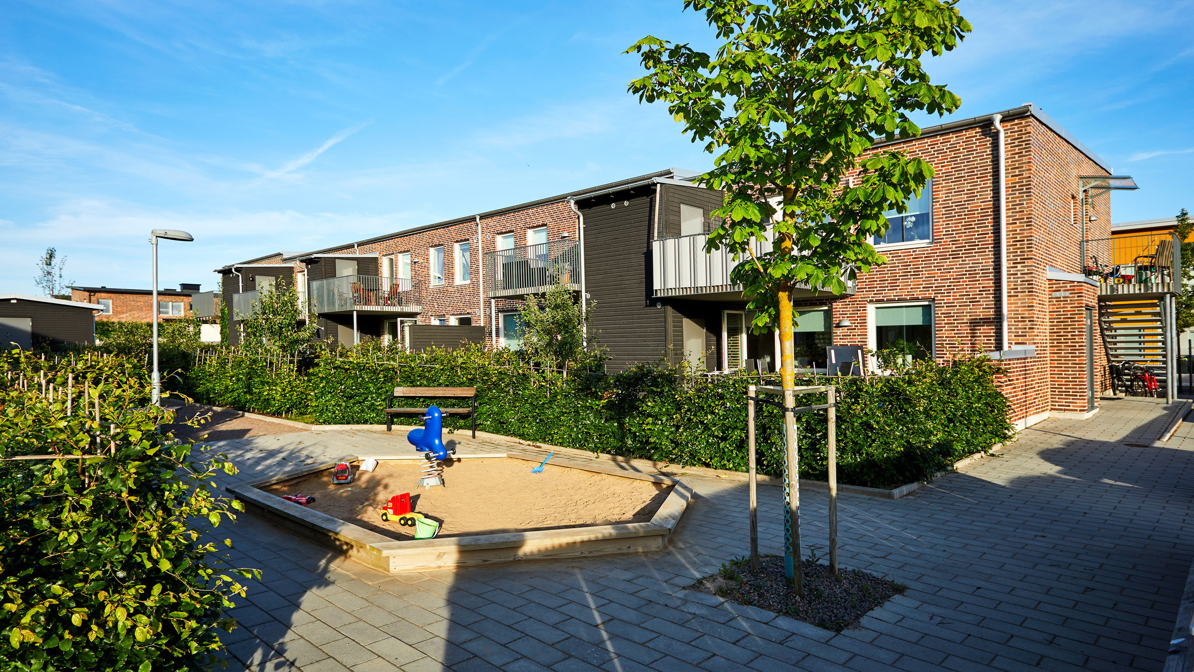 Modern bostadsområde med radhus i tegel, en lekplats och en trädkantad gångväg.