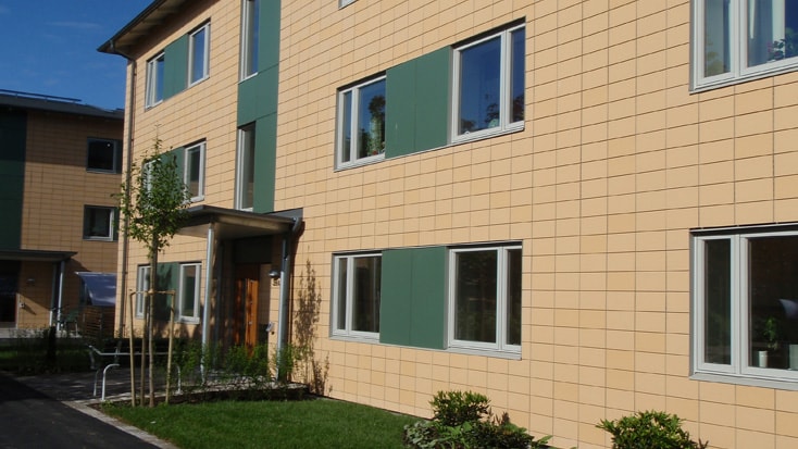 Brogården Modern lägenhetsbyggnad med beige exteriör, gröna paneler och stora fönster omgiven av grönska.