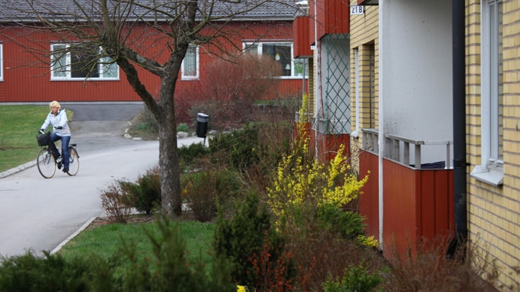 Brogården Kvinna cyklande på en stig bredvid färgglada bostadshus och gröna buskar, som förstärker stadens charm.