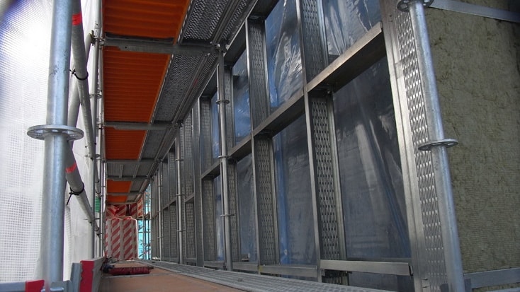 Vy över ett byggnadsställningssystem med metallramar och orangea plankor i en industriell miljö.