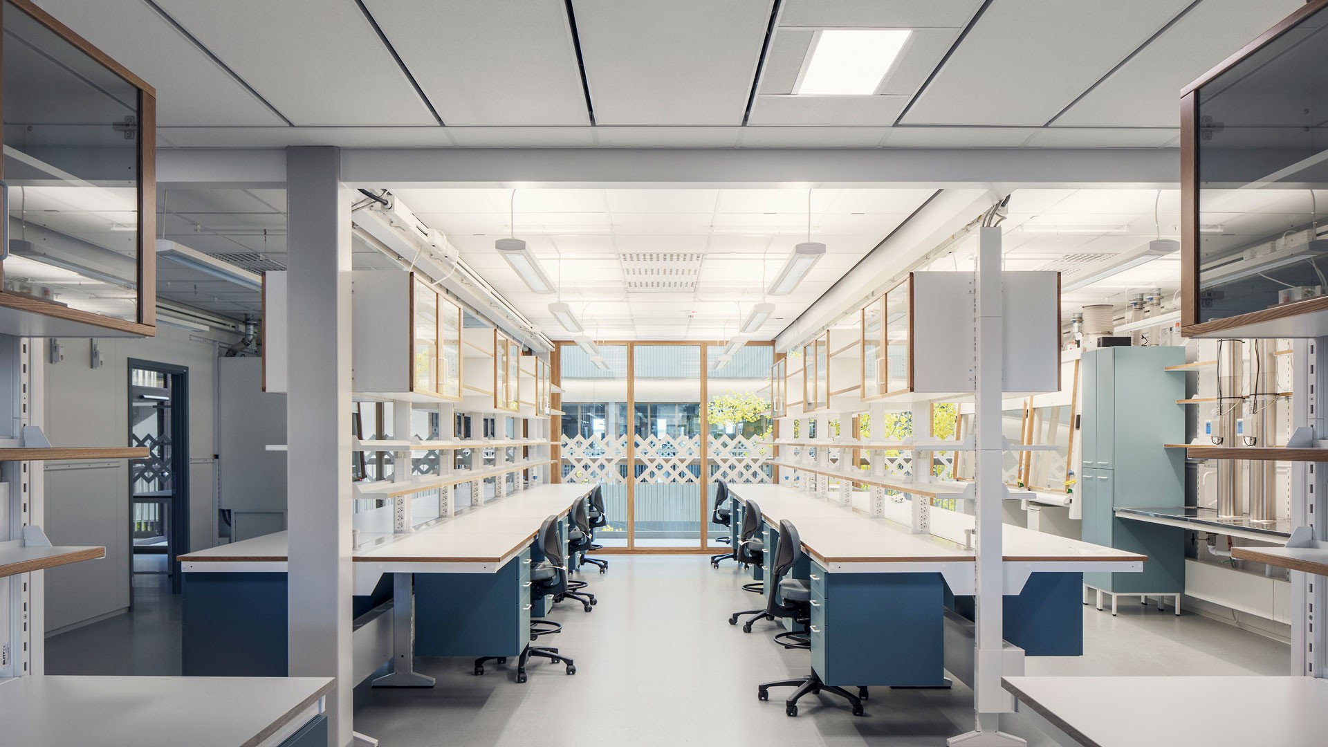 Modern laboratorieinteriör med arbetsstationer, hyllor och stolar, med ljus belysning.