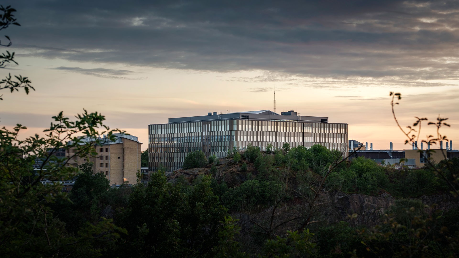 Modern byggnad på en kulle omgiven av grönska under en molnig himmel vid solnedgång.