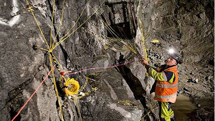 Norra Länken 11 En gruvarbetare installerar sprängladdningar på en bergvägg inne i en svagt upplyst underjordisk tunnel.