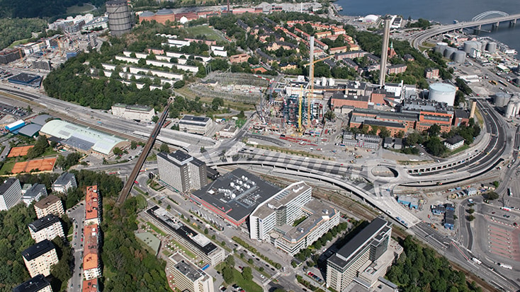 Flygvy över en urban byggarbetsplats med omgivande byggnader och infrastruktur.