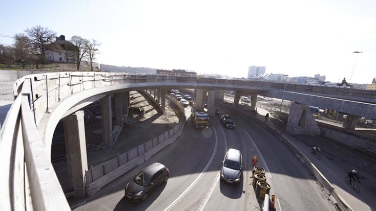 Bilar kör på en kurvig motorväg under konstruktion med synliga betongviadukter och barriärer.