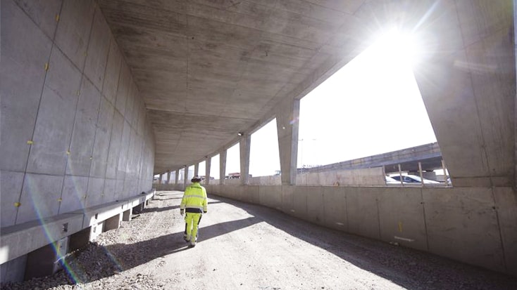 Byggnadsarbetare i varselkläder som går under en betongmotorvägsviadukt i solljus.