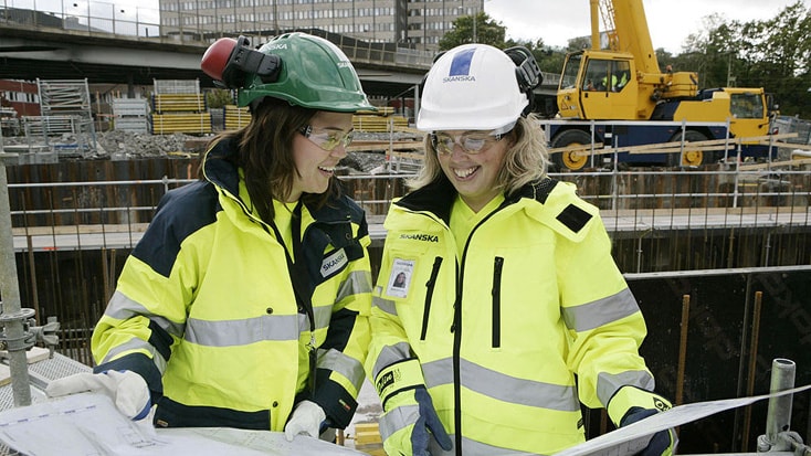 Två byggnadsarbetare i skyddsutrustning granskar planer på en byggarbetsplats med en kran i bakgrunden.