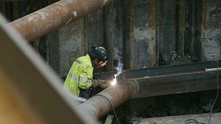 Arbetare svetsar metallbalkar på en byggarbetsplats och bär skyddsutrustning och en högsynlighetsjacka.