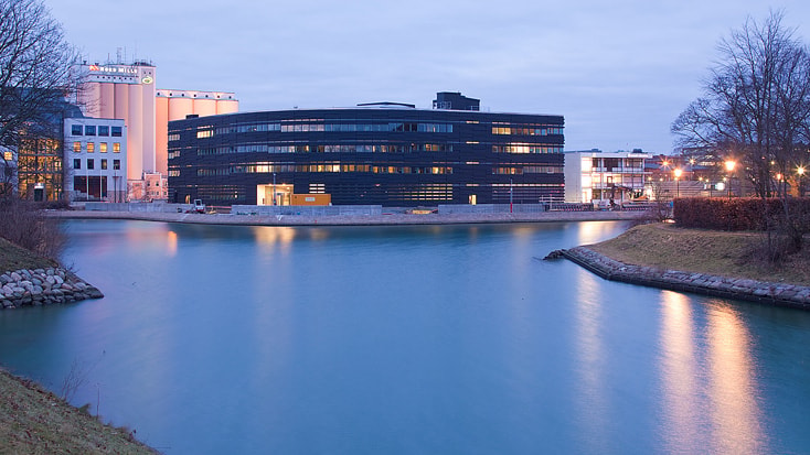 Modern byggnad vid vattnet i skymningen, omgiven av vatten och industriella strukturer i bakgrunden.