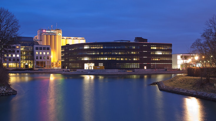 Modern byggnad vid vattnet upplyst på natten, speglas i lugnt vatten under en djupt blå himmel.