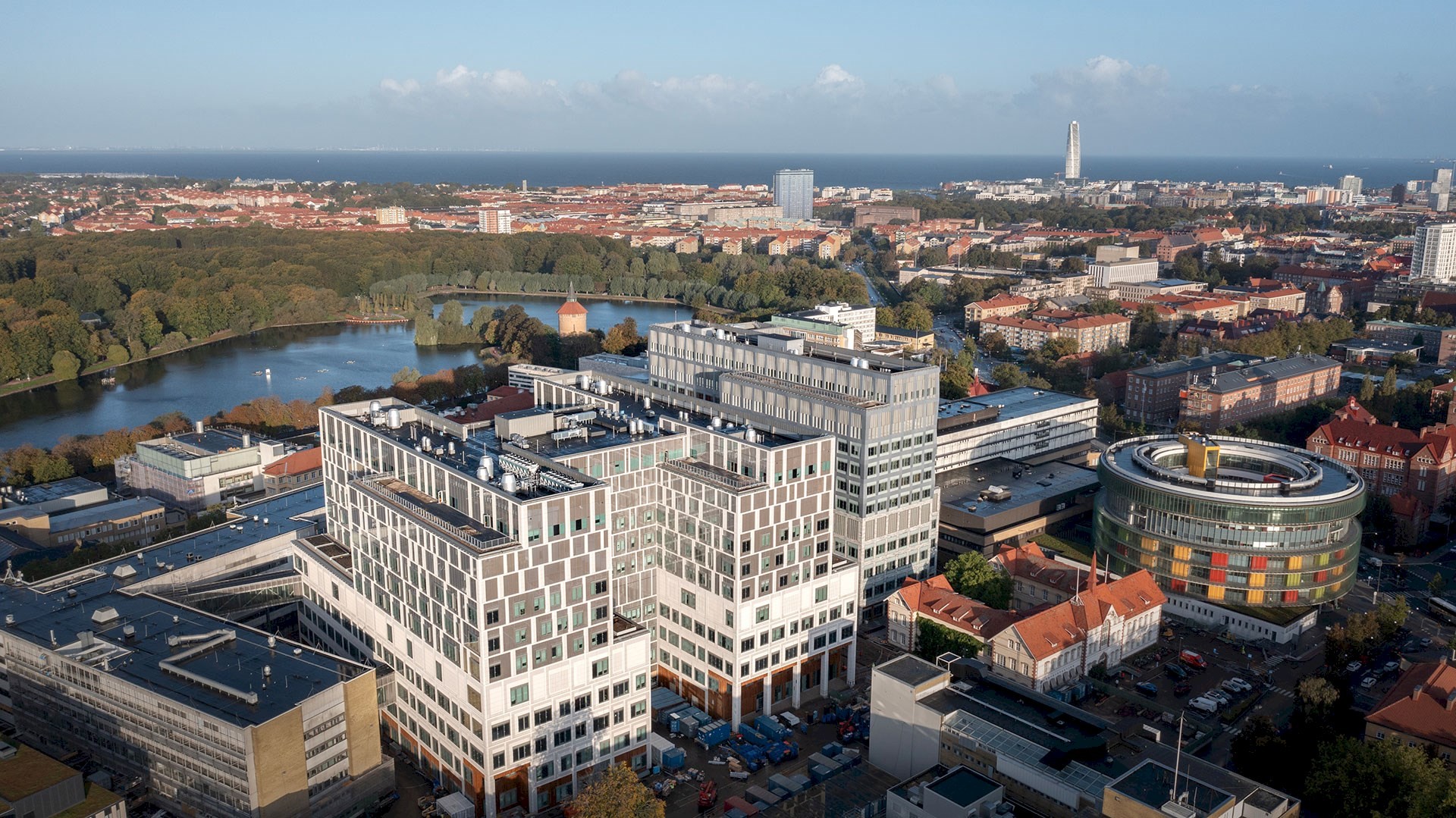 Byggnad 35, vårdbyggnaden till höger i bilden, är färdig och den 8 januari lämnades stafettpinnen vidare till kunden Region Skåne. Tre stora vita byggnader reser sig mot den lägre staden och vattnet som syns i bakgrunden.