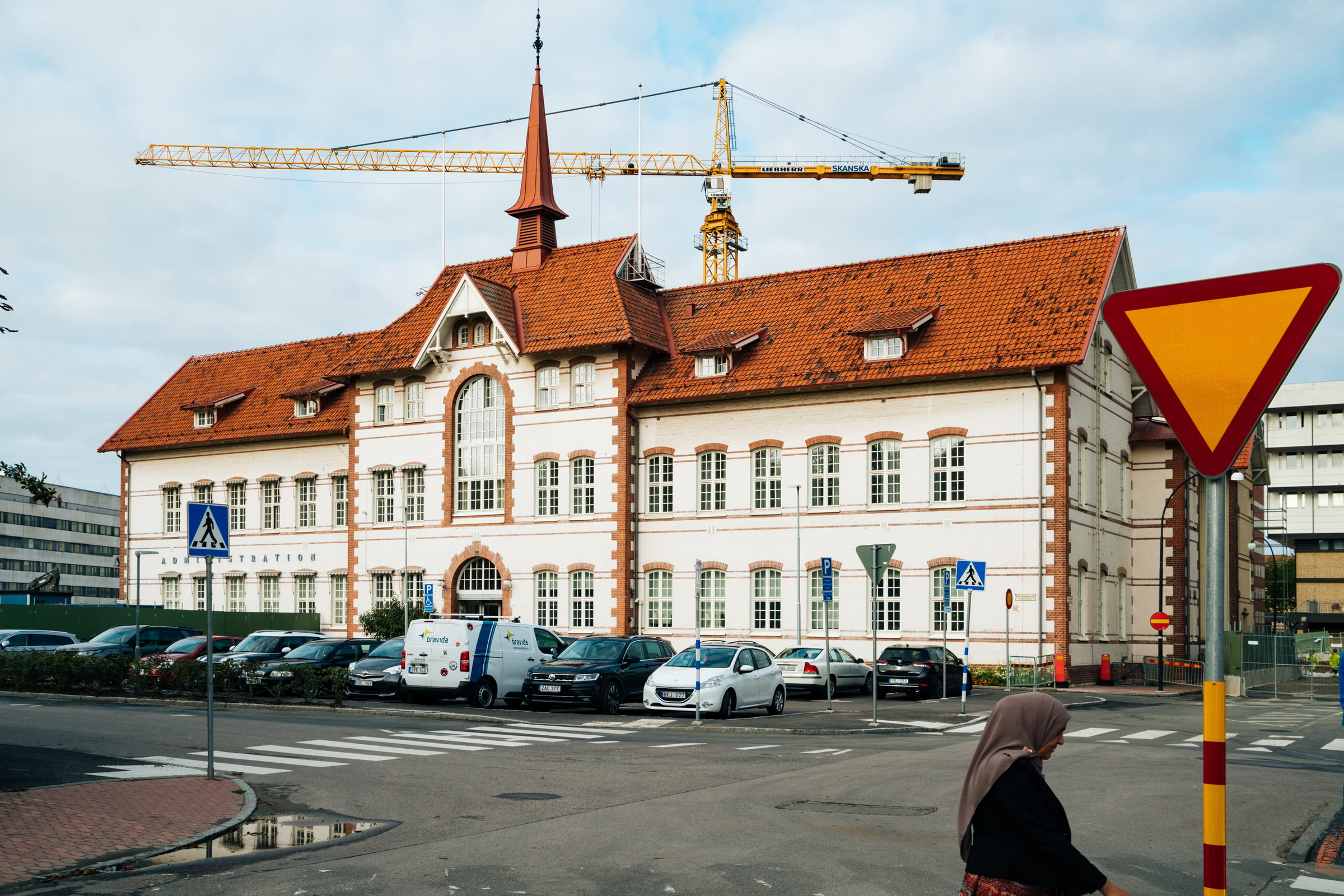 Historisk byggnad med rött tak och kran i stadsområde; fotgängare och parkerade bilar i närheten.
