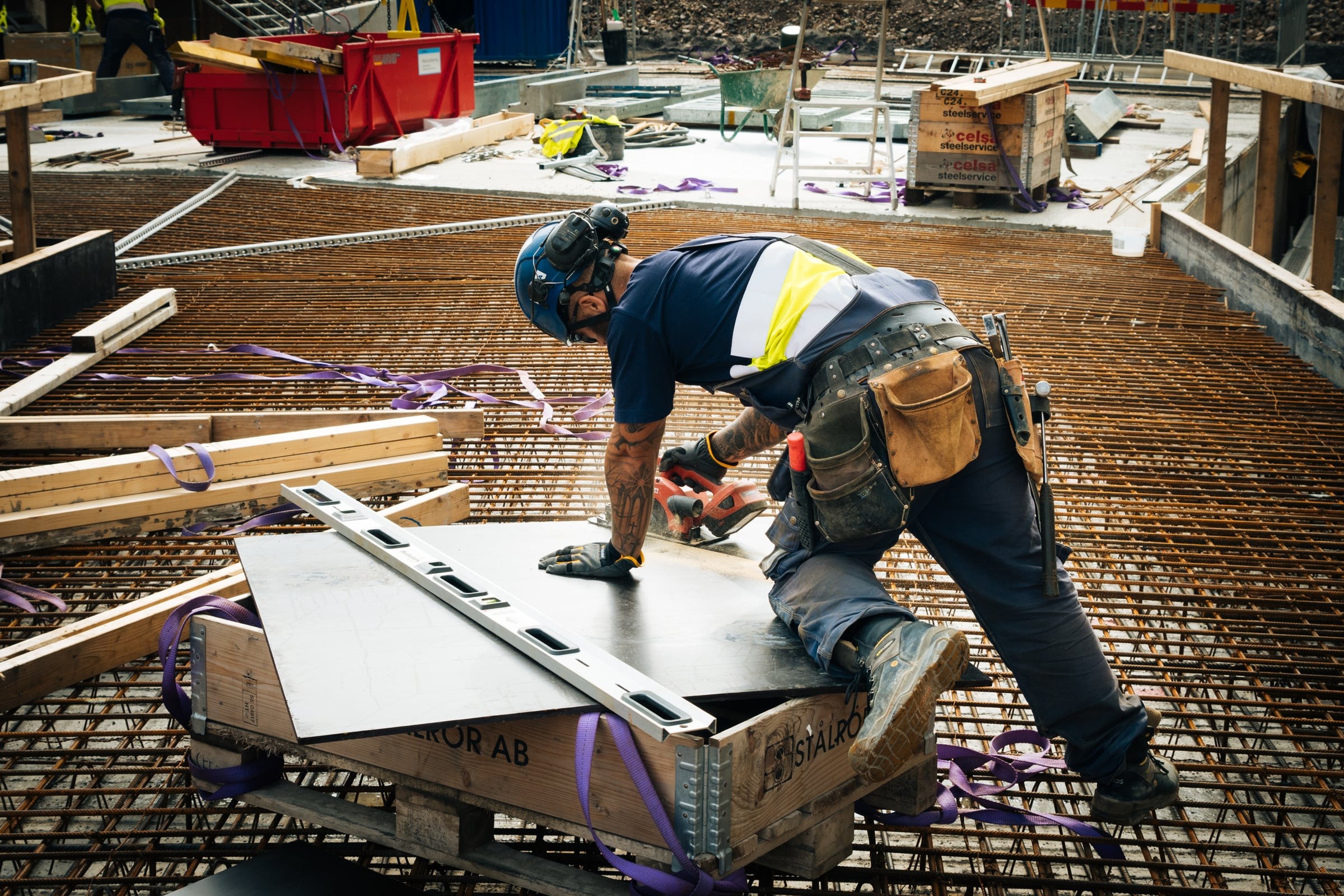 Byggarbetare skär metallplåt på plats, iförd skyddsutrustning, omgiven av verktyg och material.