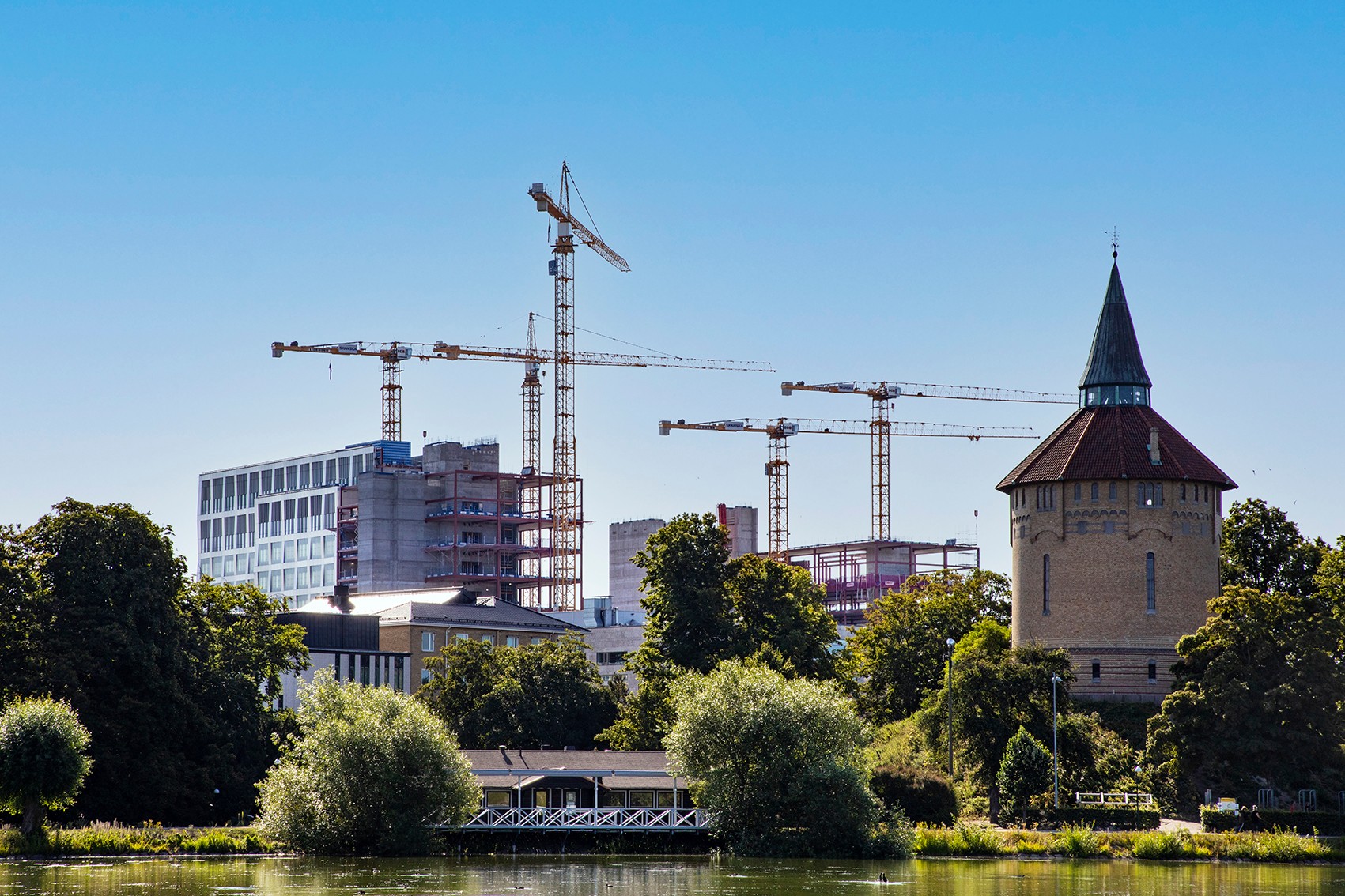 En naturskön vy av byggnadskranar bakom ett historiskt torn, omgivet av frodig grönska och vatten.
