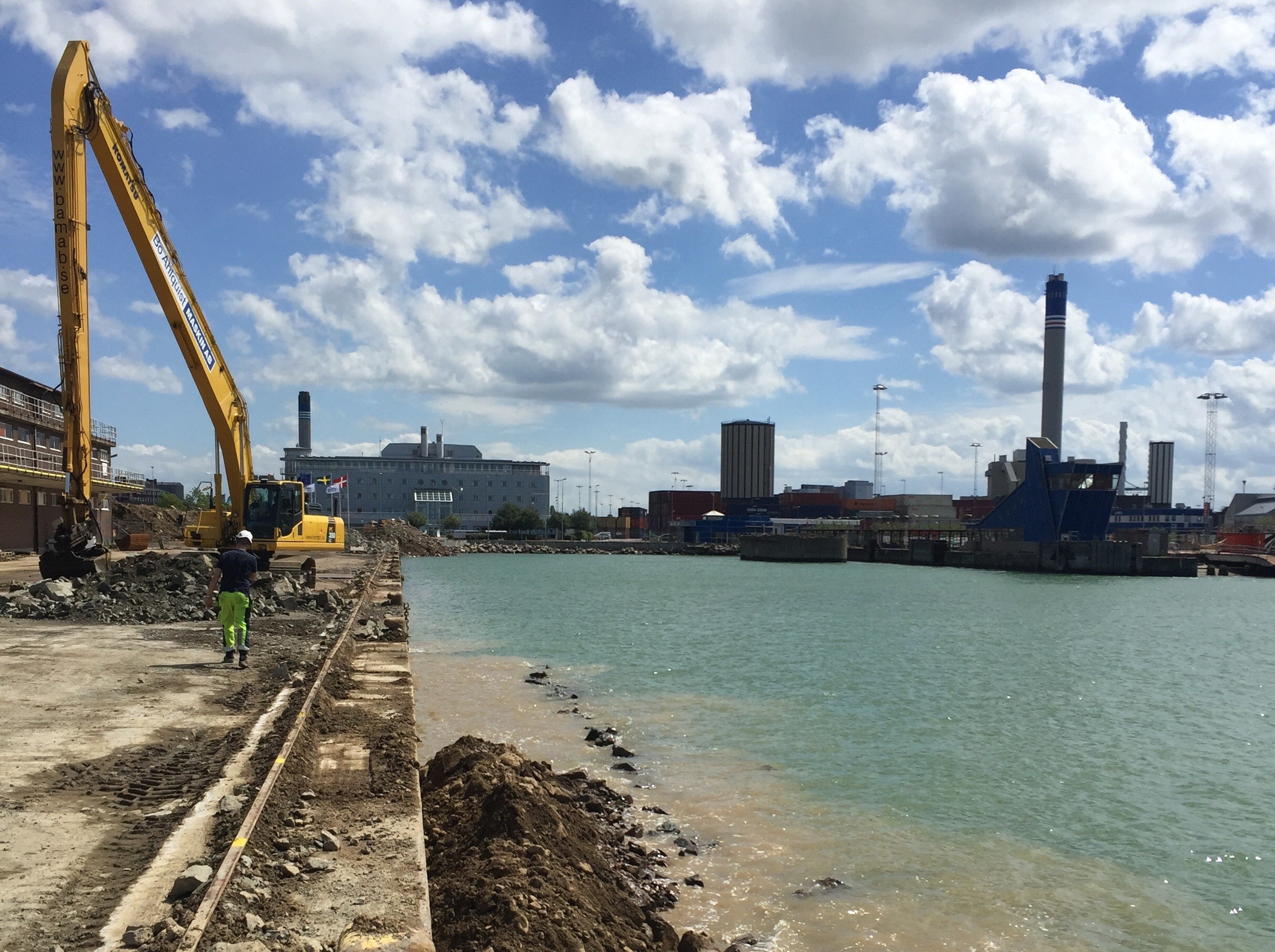 Helsingborgs stad planerar att omvandla den gamla industrihamnen till stadsbebyggelse. Byggarbetsplats vid en strandkant med en grävmaskin, industribyggnader och molnig himmel.