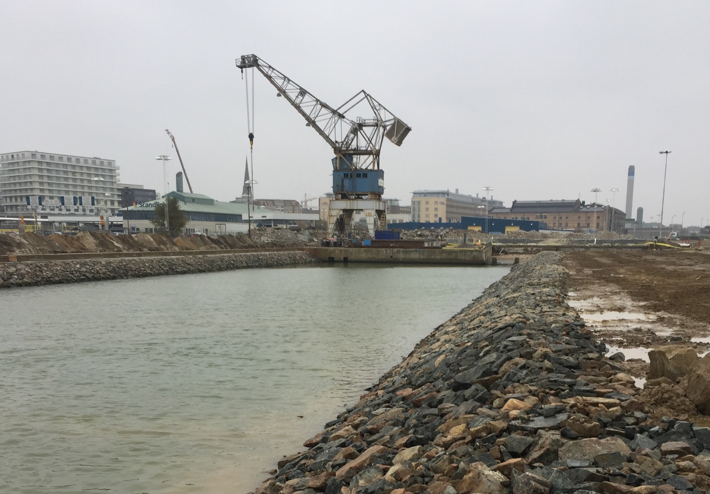 Uppdraget innefattade övertäckning av förorenade bottensediment, stabiliserande fyllnader mot gamla kajlinjer, rivningsarbete samt betongreparationer. Industriell kran vid en stenig kanal i ett varv med byggnader i bakgrunden under molniga himlar.
