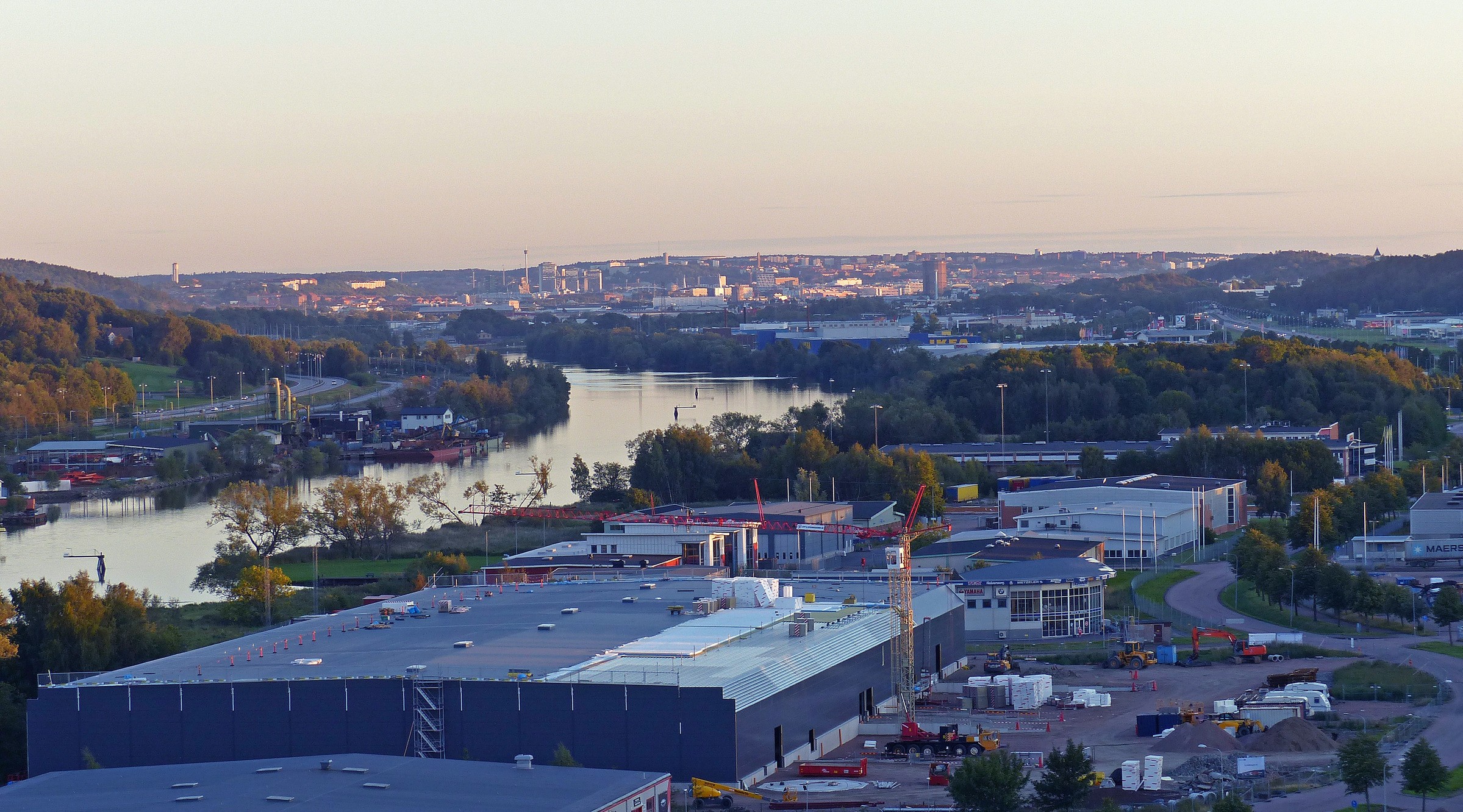 Närheten till Göta Älv har gjort att grundläggningsarbetet blev utmanande. Vi har pålat 65-70 meter men ändå inte kommit ner till fast berg. Industriområde med lager nära en flod, omgiven av grönska och stadsbild i bakgrunden.