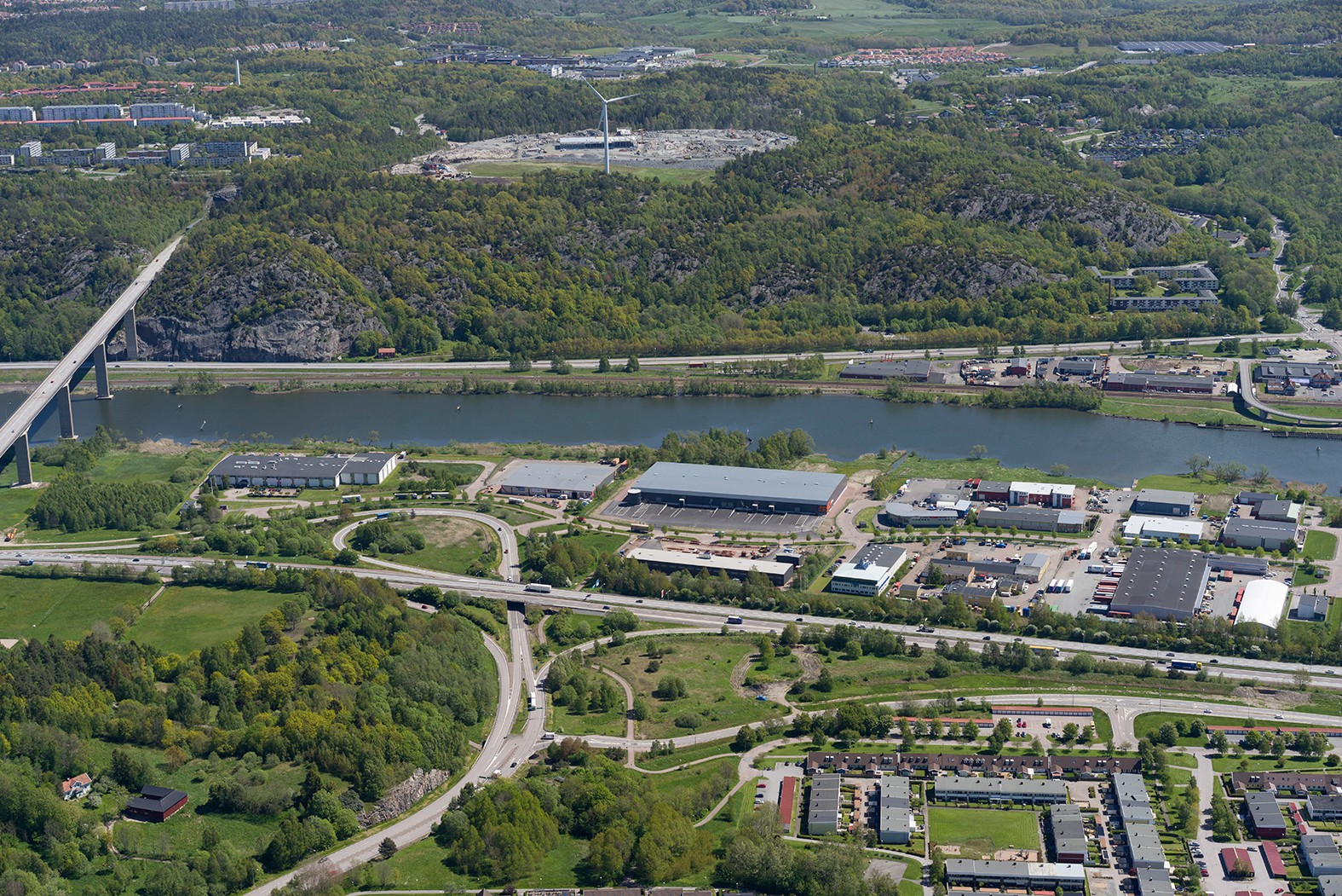 Flygvy över ett industriområde nära en flod, med omgivande grönska och avlägsna kullar.