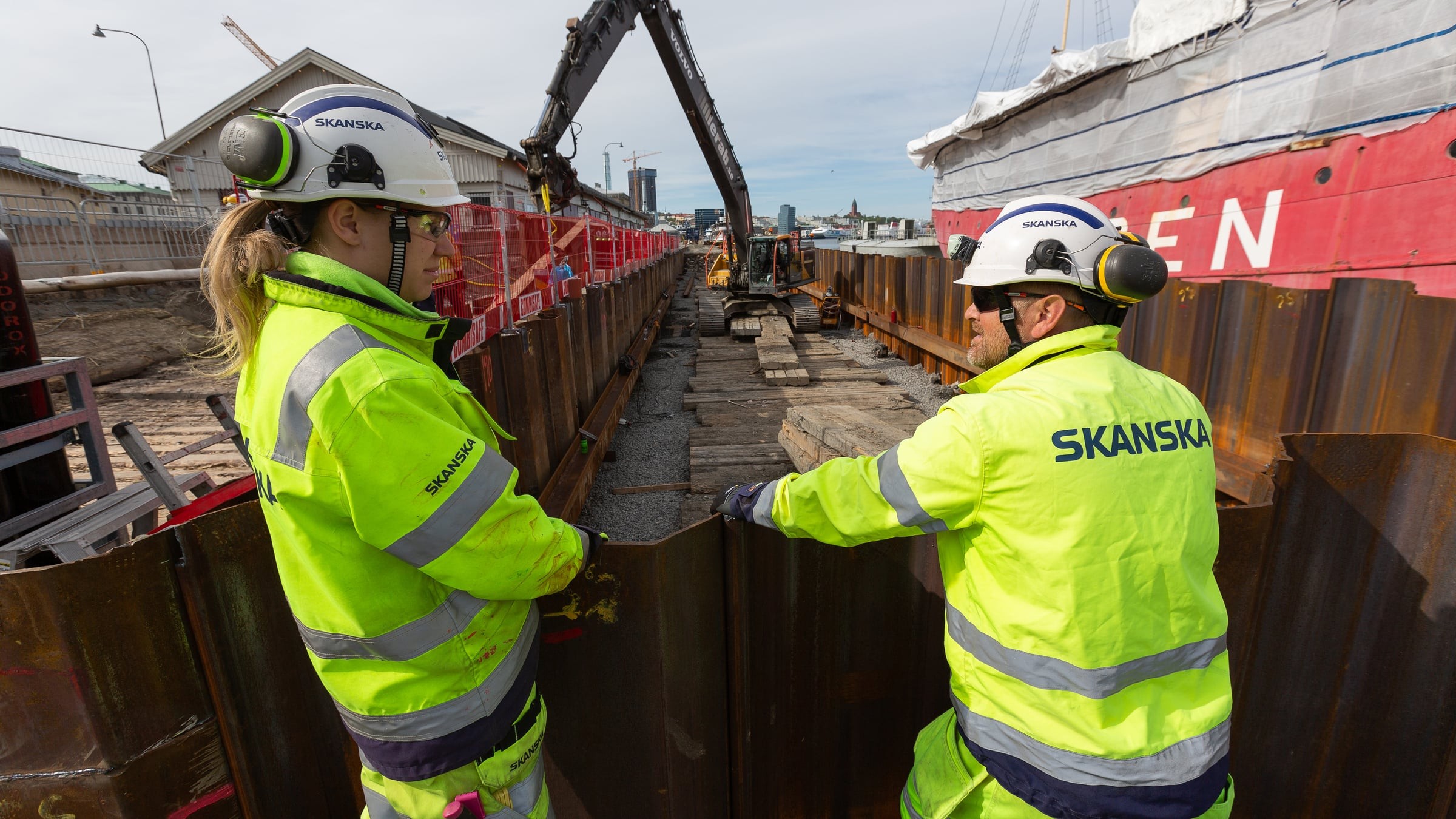 Byggnadsarbetare i Skanska-uniformer övervakar en arbetsplats med tunga maskiner och avspärrningar.