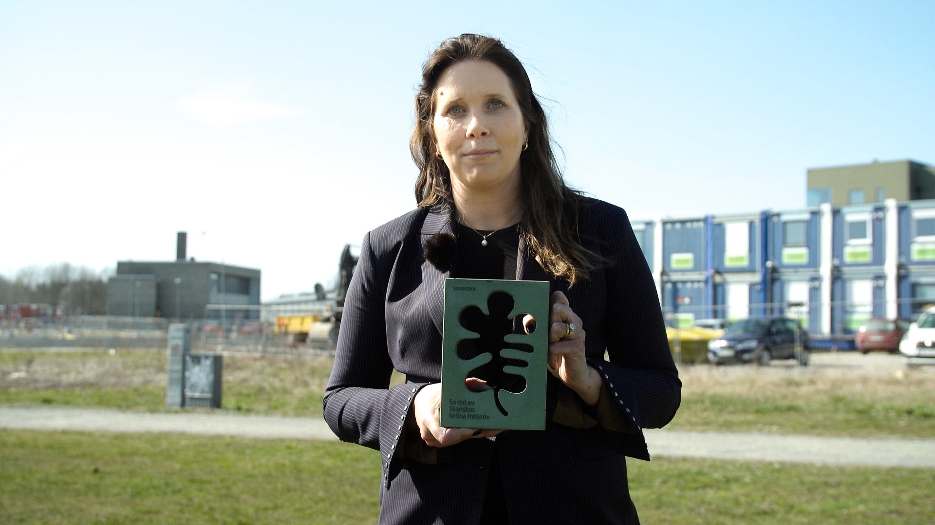 Skanska Sveriges vice vd Alexandra Laurén vid invigningen av produktionen av Parkkvarteret En person som håller en grön prisstatyett med unik design, stående utomhus nära en byggarbetsplats.