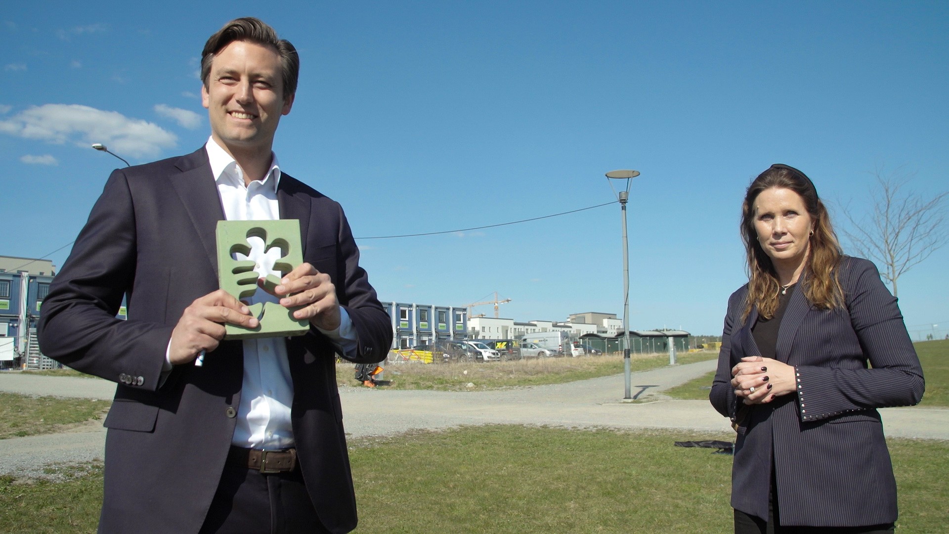 Skanska Sveriges vice vd Alexandra Laurén och Sebastian Vallgårda, transaktionschef hos fastighetsinvesteraren NREP Två personer står utomhus, en håller i en grön bok, med byggnader och klar himmel i bakgrunden.