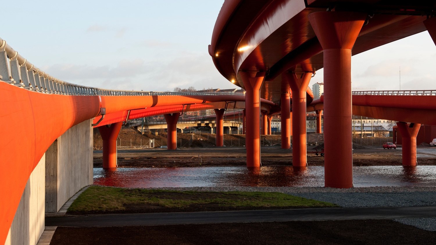 Partihallsförbindelsen Röde Orm Röd upphöjd gångbro med böjda gångvägar och stödjande pelare över en vattenmassa.