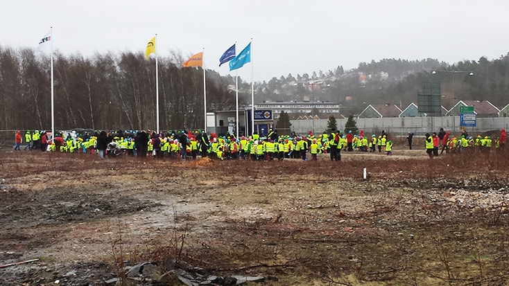 En stor grupp iklädd varseljackor samlas nära flaggstänger på ett öppet, lerigt fält.