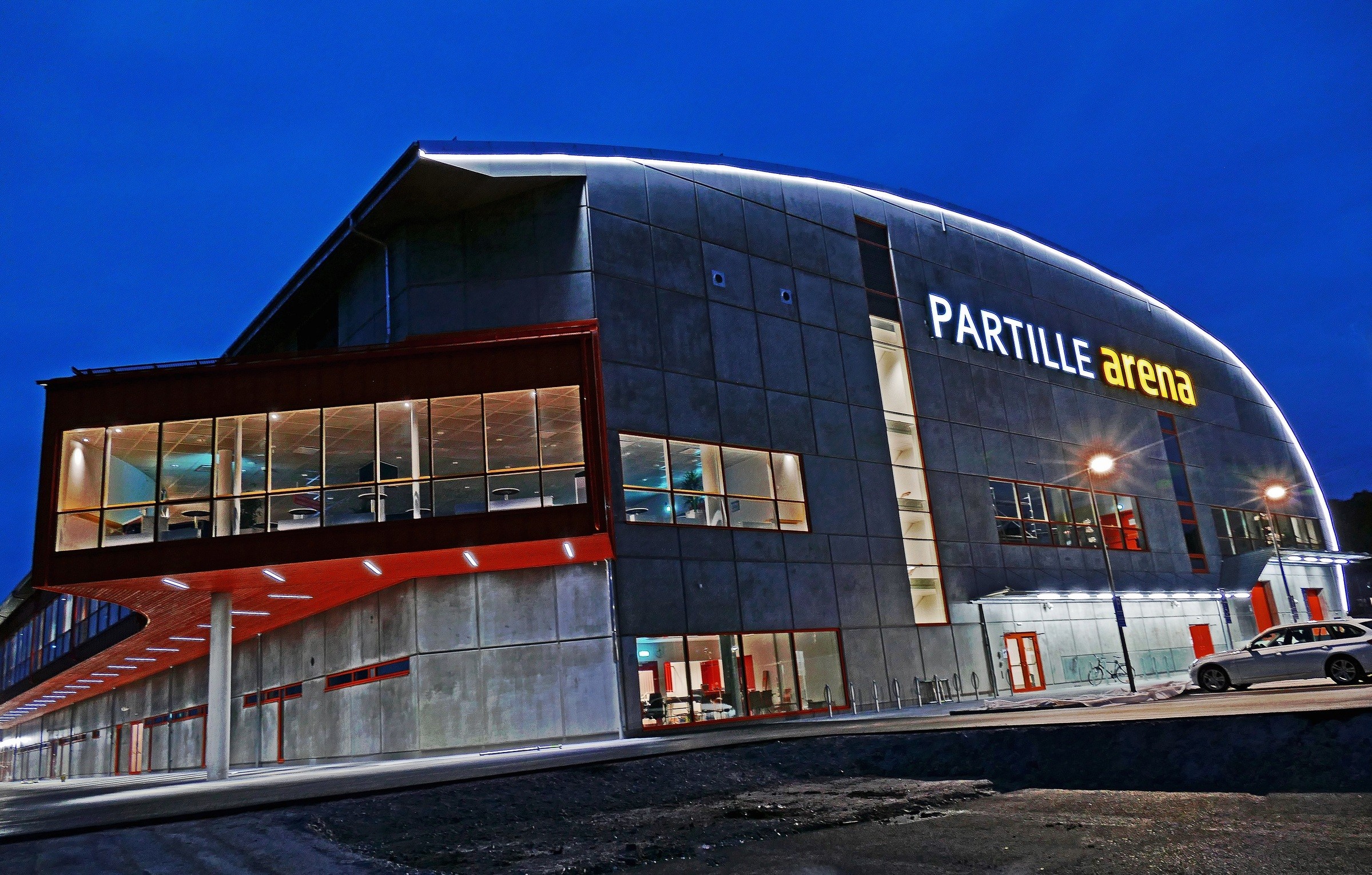 Modern Partille Arena med upplyst fasad och stora glasfönster mot en skymningshimmel.