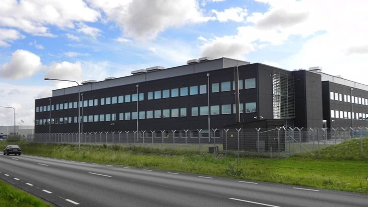 Modern industribyggnad med stora fönster, omgiven av ett säkerhetsstängsel längs en väg.