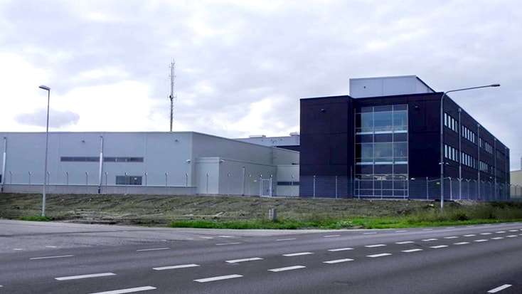 Modern industribyggnad med stora fönster och inhägnad perimeter längs en bred väg.