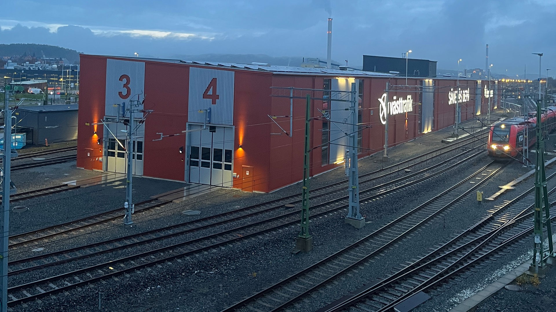 2021 fick Skanska i uppdrag av Västtrafik och Jernhusen att bygga den nya tågdepån. Modern järnvägsunderhållsanläggning upplyst i skymningen med järnvägsspår och ett tåg i sikte.