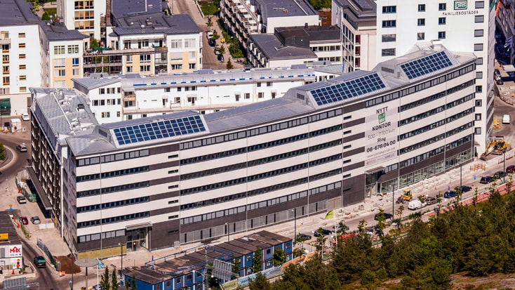 Modern kontorsbyggnad med solpaneler på taket, omgiven av urban infrastruktur.